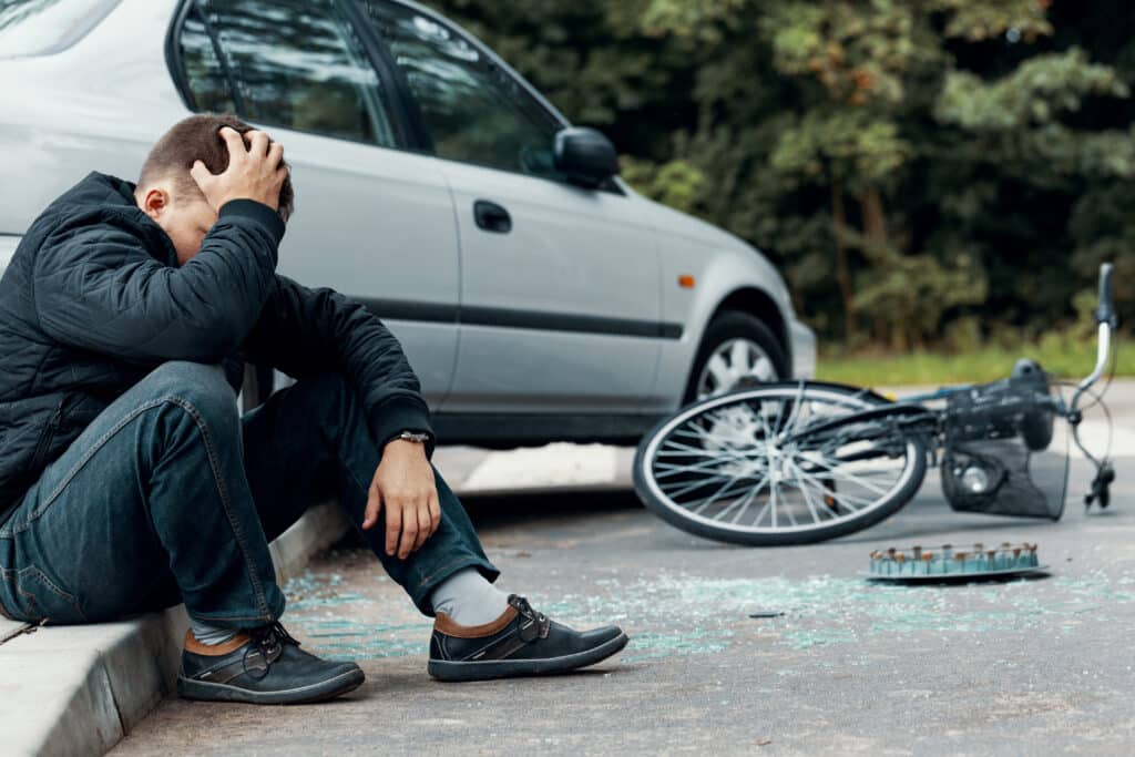 Worried biker holding his head and sitting on a pavement next to a car and bike crash. Concussion/mTBI